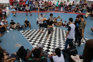 dançarinos dançam break num palco xadrez preto e branco, enquanto o público acompanha em arquibancadas circundando o tablado