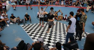 dançarinos dançam break num palco xadrez preto e branco, enquanto o público acompanha em arquibancadas circundando o tablado