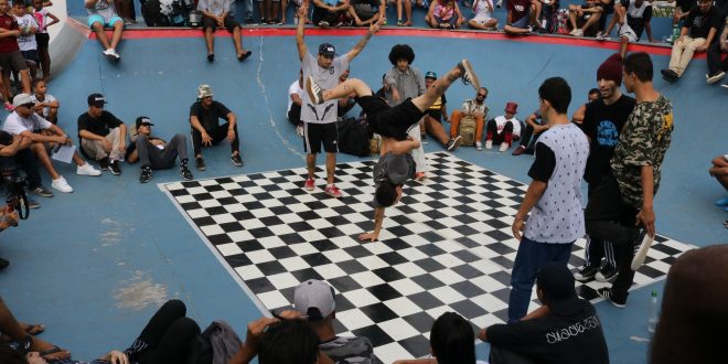 dançarinos dançam break num palco xadrez preto e branco, enquanto o público acompanha em arquibancadas circundando o tablado