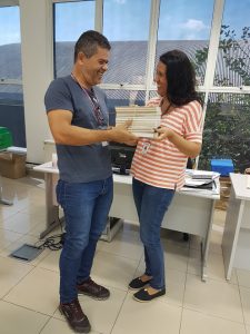 um professor entrega uma pilha de livros para outra professora enquanto ambos sorriem no interior de uma escola de Barueri