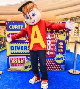 Pessoa fantasiada de personagem da Gloob posta de braços abertos para a câmera diante de instalação para crianças