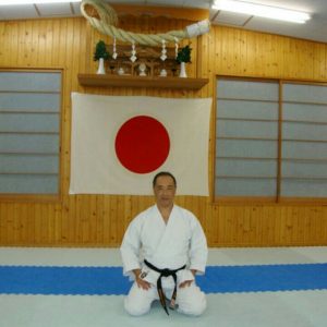 Ruy Koike ajoelhado de kimono branco diante de uma bandeira do Japão estendida na parede