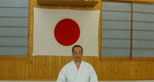Ruy Koike ajoelhado de kimono branco diante de uma bandeira do Japão estendida na parede