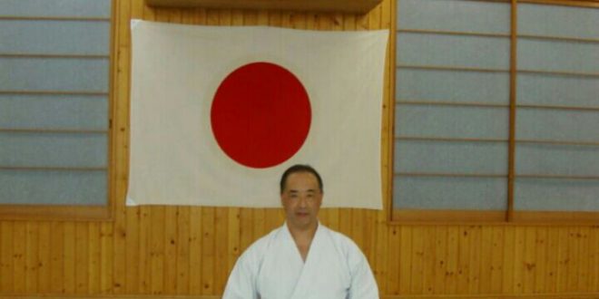 Ruy Koike ajoelhado de kimono branco diante de uma bandeira do Japão estendida na parede