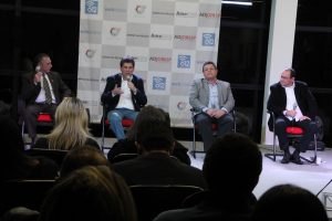 Claudio Piteri e demais candidatos sentado no palco do debate, com as cabeças de alguns membros da plateia visíveis