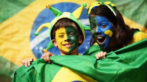 crianças com os rostos pintados para torcer pelo Brasil, com uma bandeira brasileira ao fundo