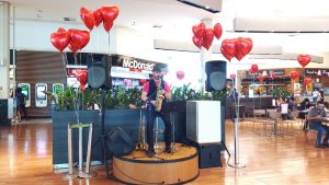 um músico se apresentando no palco da praça de alimentação do Parque Shopping Barueri com balões de corações dispostos em volta durante o Barueri Music especial de Dia dos Namorados