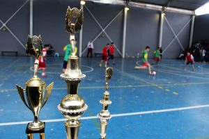 três troféus em primeiro plano, com uma quadra poliesportiva em segundo plano com uma partida de futsal acontecendo