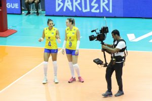 duas jogadoras da seleção brasileira de vôlei sendo filmadas na quadra por um cinegrafista