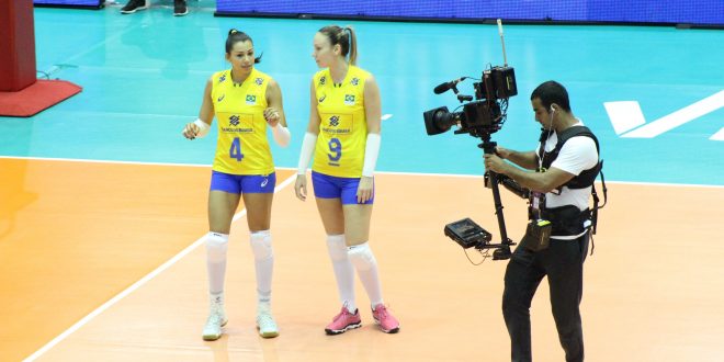 duas jogadoras da seleção brasileira de vôlei sendo filmadas na quadra por um cinegrafista