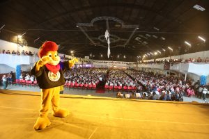 leão do proerd no palco, com milhares de pessoas ao fundo, incluindo uma banda militar da polícia militar