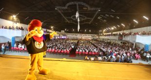 leão do proerd no palco, com milhares de pessoas ao fundo, incluindo uma banda militar da polícia militar
