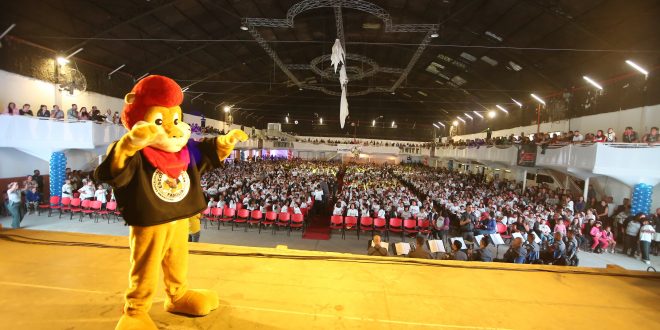 leão do proerd no palco, com milhares de pessoas ao fundo, incluindo uma banda militar da polícia militar