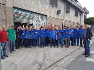 metalúrgicos da Eltra reunidos diante de um galpão abandonado com os braços erguidos