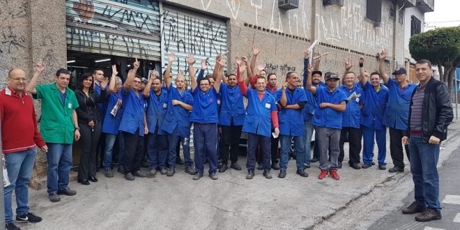 metalúrgicos da Eltra reunidos diante de um galpão abandonado com os braços erguidos