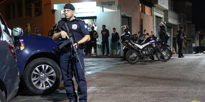 guarda civil de Barueri armado com fuzil e parado ao lado de uma viatura, patrulhando uma rua de Barueri com jovens abordados ao fundo