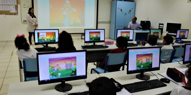 alunos sentados diante de computadores enquanto uma professora leciona ao fundo