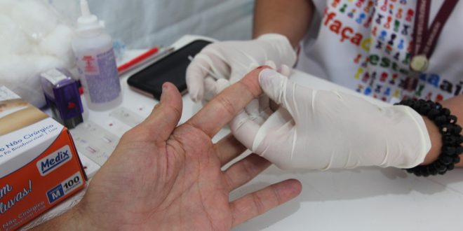 close da mão de uma pessoa que está realizando exame com coleta de gota de sangue, com um par de mãos com luvas manuseando-a