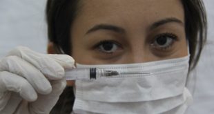 médica com máscara medicinal segurando uma seringa à frente de seu rosto enquanto olha para a câmera