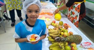 aluna posa para foto com pratinho cheio de frutas ao lado de mesa com várias frutas