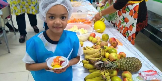 aluna posa para foto com pratinho cheio de frutas ao lado de mesa com várias frutas