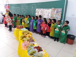 alunos e professores alinhados na parede atrás de uma mesa cheia de frutas