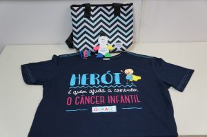 produtos que serão vendidos em prol do Graacc dispostos em uma mesa. Há uma camiseta preta escrito 'Herói é quem ajuda a combater o câncer infantil' e a palavra Graacc logo abaixo; uma bolsa térmica, um boneco com cinco marca textos fincados nos locais onde estariam sua cabeça e membros; e um chaveiro com a palavra 'fé'
