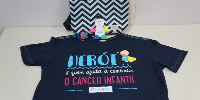 produtos que serão vendidos em prol do Graacc dispostos em uma mesa. Há uma camiseta preta escrito 'Herói é quem ajuda a combater o câncer infantil' e a palavra Graacc logo abaixo; uma bolsa térmica, um boneco com cinco marca textos fincados nos locais onde estariam sua cabeça e membros; e um chaveiro com a palavra 'fé'