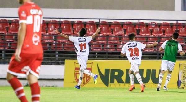 jogadores do Oeste celebram um gol com um jogador do CRB caminhando em primeiro plano