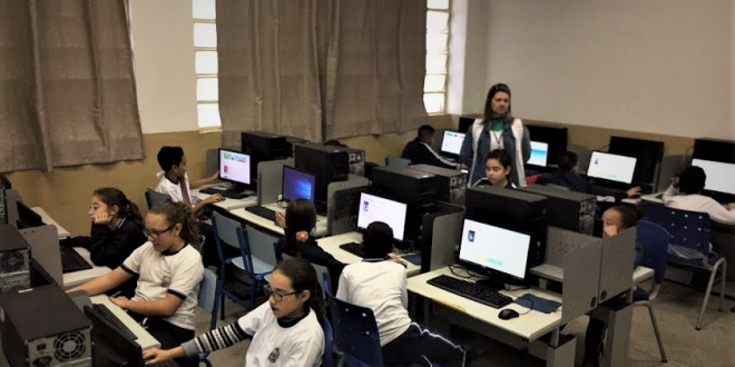 um laboratório de informática com uma turma de alunos sendo orientada por uma professora