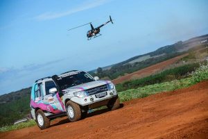 mitsubishi pilotada por Sandra se desloca numa pista de terra com um helicóptero ao fundo