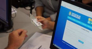 um servidor recebendo documentos de um munícipe com um computator entre eles; somente as mãos deles são vistas