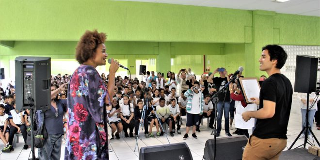 luciana melo se apresentando no palco da Emef Sidney Santucci com um músico, com o público acompanhando atrás