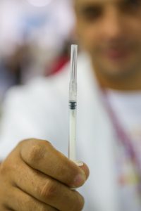 uma vacina contra meningite sendo segurada por um médico que aparece ao fundo, desfocado