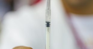 uma vacina contra meningite sendo segurada por um médico que aparece ao fundo, desfocado